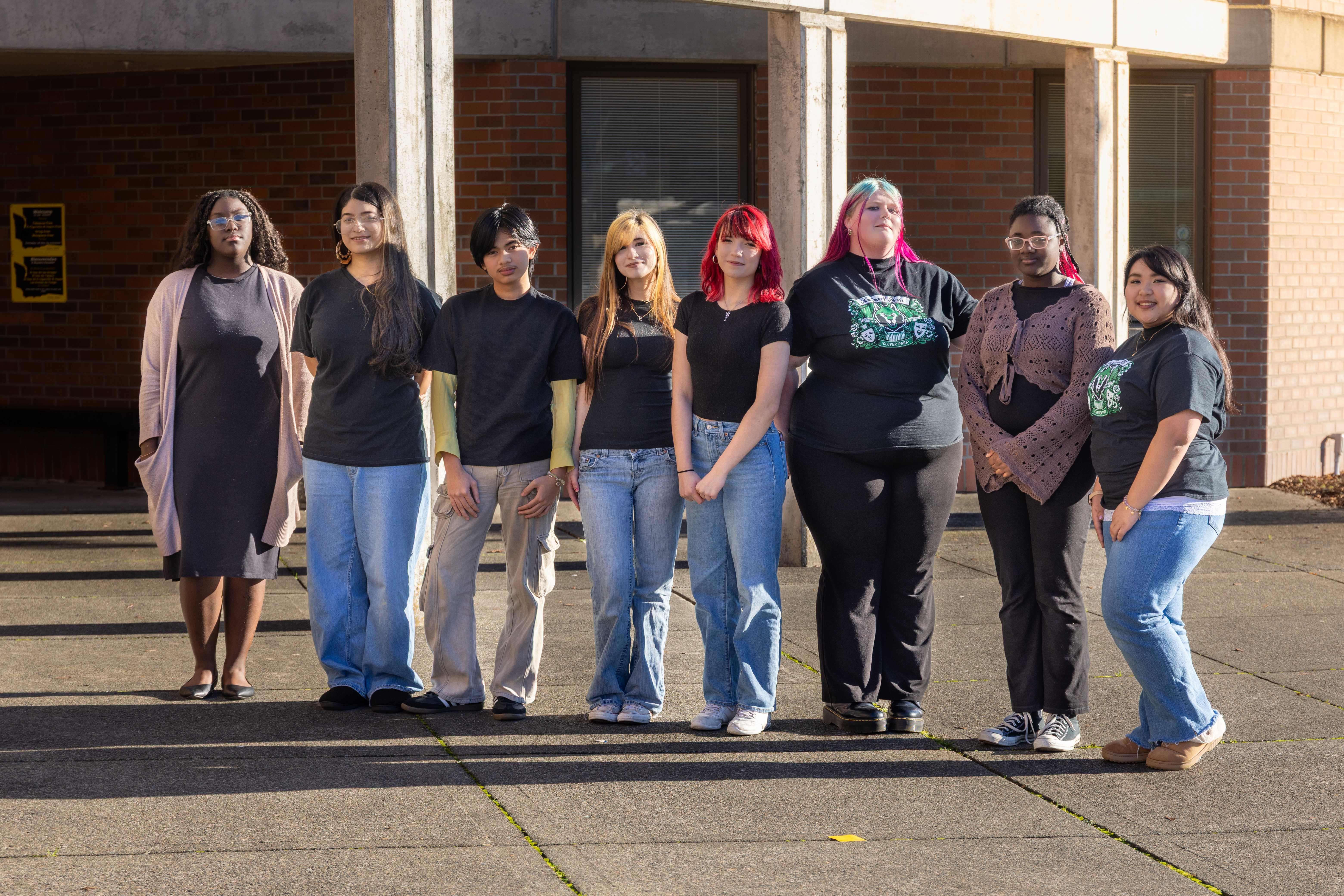 Clover Park High School drama students dressed in black shirts and jeans standing in a row and smiling outside the entrance of the high school.